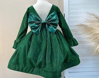 Op de afbeelding: Een groene jurk met lange mouwen en een grote strik op de rug. De jurk heeft een V-hals en een volle rok. De stof heeft een subtiele glans. De jurk wordt op een mannequin gepresenteerd.