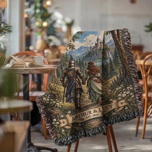 Pu&ograve; includere: Una coperta tessuta con una scena fantasy che ritrae un cavaliere in armatura e una persona con un abito lungo che camminano verso un castello tra le montagne. Il design include uno stendardo con il testo ARTHUR & LUCY EST. 2026 e bordi sfrangiati.