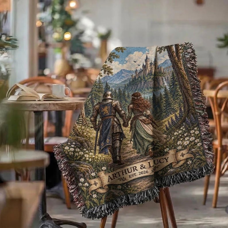 Pu&ograve; includere: Una coperta tessuta con una scena fantasy che ritrae un cavaliere in armatura e una persona con un abito lungo che camminano verso un castello tra le montagne. Il design include uno stendardo con il testo ARTHUR & LUCY EST. 2026 e bordi sfrangiati.