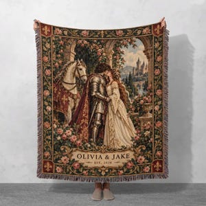 Peut inclure: Un plaid en tapisserie tissée représentant un chevalier en armure et une personne en robe s'embrassant près d'un cheval blanc. La scène inclut un château, des arches en pierre et des roses roses. Le texte indique OLIVIA & JAKE EST. 2026.