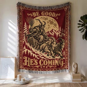 Pode incluir: Uma manta retangular tecida em vermelho escuro e creme com o gráfico de uma figura com chifres num trenó puxado por duas cabras. O texto BE GOOD e HE'S COMING está impresso numa fonte decorativa. A manta tem uma borda com franjas.