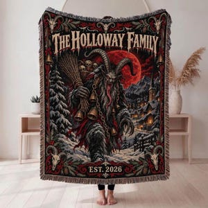 May include: A woven throw blanket featuring a dark, detailed illustration of a horned creature with a long tongue, carrying a bundle of sticks and bells. The text reads THE HOLLOWAY FAMILY and EST. 2026, set against a snowy mountain village backdrop.