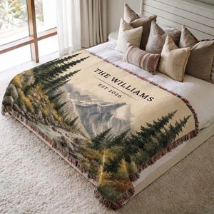May include: A woven throw blanket featuring a landscape of pine trees and mountains in shades of green, brown, and cream. The text THE WILLIAMS EST 2026 is printed in black on the upper right section. The blanket has a fringed edge on all sides.