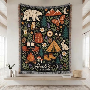 May include: A black woven throw blanket featuring a camping-themed pattern with a bear, mountains, butterfly, owl, lantern, backpack, compass, tent, rabbit, campfire, and hiking boots. The text reads Alex & Jamie EST. 2026 with floral accents.
