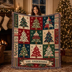 May include: A woven throw blanket featuring a grid of nine Christmas trees in various colors and patterns, including red, green, and blue. The bottom border displays a banner with the text THE ANDERSONS, surrounded by holly and pine.