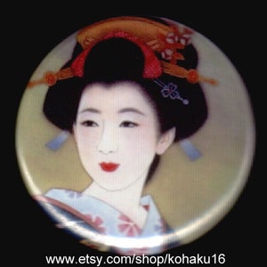 May include: A round button featuring a colorful illustration of a woman in traditional Japanese clothing. She has dark hair with a red and gold headband and is wearing a white and pink kimono. The background is a soft yellow.