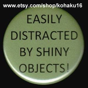 May include: A green round button with the text "EASILY DISTRACTED BY SHINY OBJECTS!" in black.