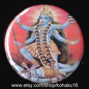 May include: A round button with a colorful image of a Hindu goddess with multiple arms, wearing a blue outfit and a gold crown. The goddess is standing on a figure with a snake wrapped around it. The background is red.