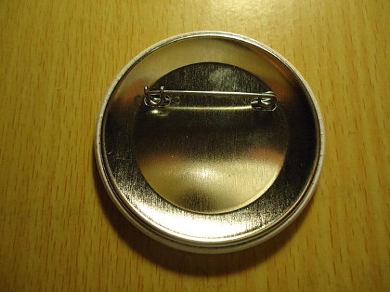 May include: The back of a white button with a silver pin and a metal clasp. The button is round and has a smooth surface.
