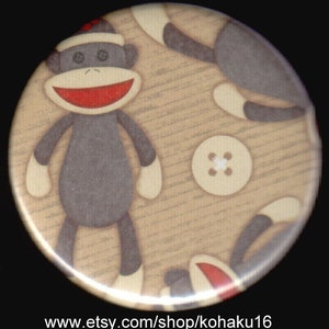 May include: A round button with a beige background featuring a repeating pattern of gray sock monkeys wearing red Santa hats. The button also features a white button with four holes in the center.