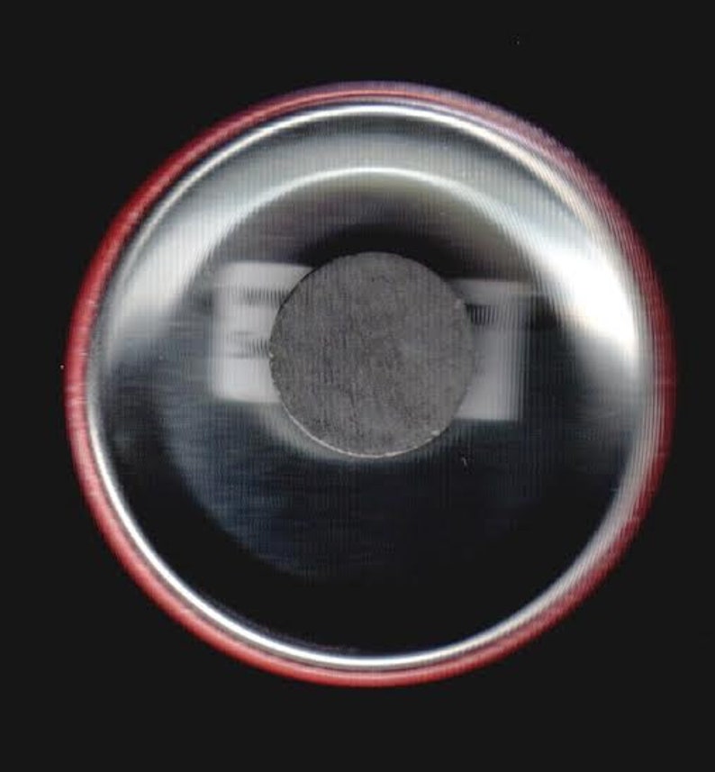Puede incluir: Un bot&oacute;n redondo de color plateado con un borde rojo. El bot&oacute;n tiene un im&aacute;n circular negro en el centro.