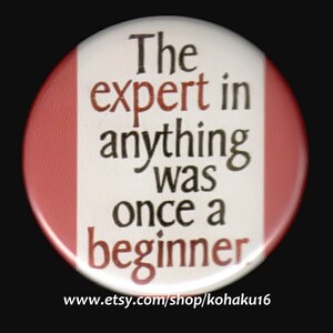 May include: A white button with red trim features the text "The expert in anything was once a beginner". The text is in black with a brown outline. The button has a website address printed on the bottom: "www.etsy.com/shop/kohaku16".