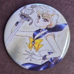 May include: A round button featuring a manga-style illustration of a character holding a sword. The character wears a white and blue uniform with a yellow bow. The button has a glossy finish and a light beige background.