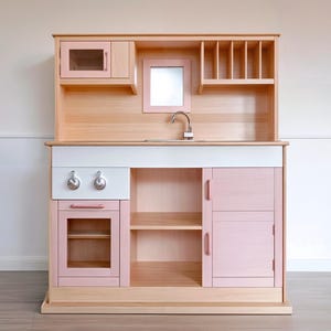 May include: A wooden toy kitchen in light pink and natural wood. It has a microwave, oven, sink, and storage shelves. The knobs and tap are silver. A fun and engaging toy for children.