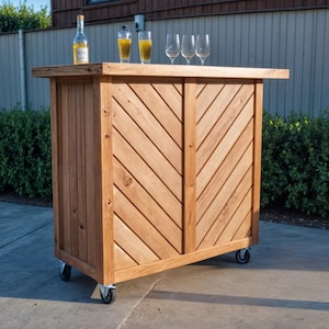 Puede incluir: Una barra de exterior de madera sobre ruedas con un patrón de espiga en la parte delantera. La barra tiene una encimera con una botella y dos vasos de líquido, más tres copas de vino vacías. La barra es de madera marrón claro.