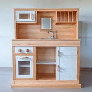 Könnte beinhalten: Eine Spielzeugküche aus Holz mit hellbraunem Finish. Sie verfügt über eine Mikrowelle, ein Waschbecken und Schränke mit weißen und hellbraunen Türen. Die Spielzeugküche umfasst einen Wasserhahn, einen Spiegel und ein Regal.