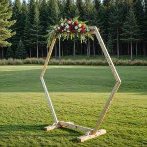 May include: Wooden wedding arch with a geometric design, adorned with a floral arrangement of red and white flowers and greenery. The arch is set on a green lawn with a backdrop of tall pine trees.