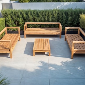 Puede incluir: Conjunto de muebles de exterior de madera clara, compuesto por un sofá de dos plazas, dos bancos individuales y una pequeña mesa de centro rectangular. El mobiliario está dispuesto sobre un patio embaldosado gris claro, rodeado de vegetación.