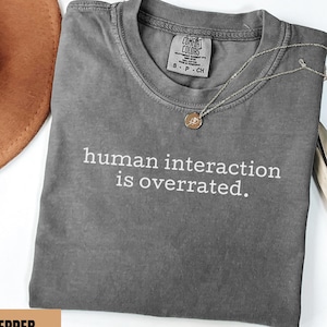 May include: A gray t-shirt with the phrase "human interaction is overrated" in white text. The shirt has a crew neck and short sleeves. A gold necklace with a small pendant is on the shirt. A brown hat is in the background.