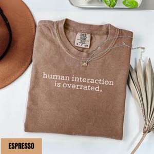 May include: Espresso-colored t-shirt with the text "human interaction is overrated." A silver necklace with a pendant rests on the shirt. The shirt is folded on a white surface, with a brown hat and dried palm leaves in the background.
