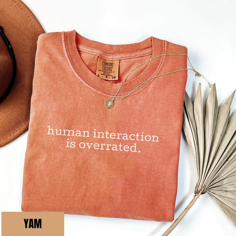 May include: A coral-colored t-shirt with the phrase "human interaction is overrated" in white lettering. A gold necklace with a pendant rests on the shirt. A brown hat and dried palm leaf are in the background.