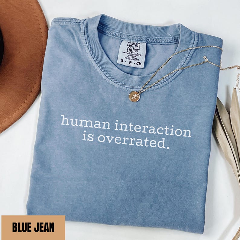 May include: A blue jean colored t-shirt with the text "human interaction is overrated" in white. The shirt has a crew neck and short sleeves. A gold necklace with a pendant is draped across the shirt.