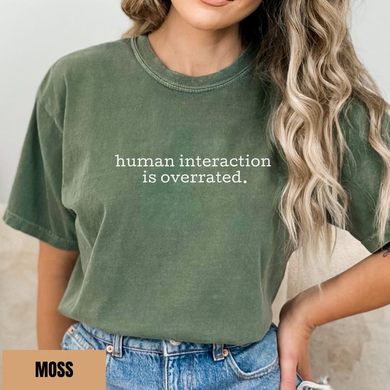 May include: Moss green t-shirt with the text "human interaction is overrated." The shirt has a crew neck and short sleeves. The word "MOSS" is displayed in a tan rectangle at the bottom of the image. The shirt is made of a soft material.