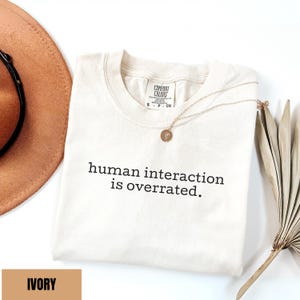 May include: Ivory-colored Comfort Colors t-shirt with the text "human interaction is overrated." A gold necklace with a pendant rests on the shirt. A brown hat and dried plant are in the background.