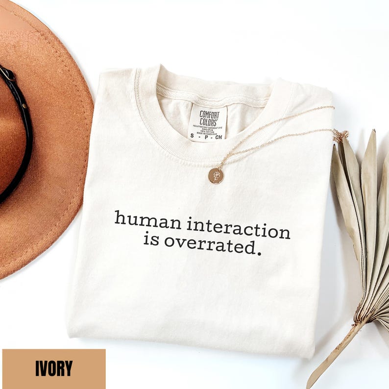 May include: Ivory-colored Comfort Colors t-shirt with the text "human interaction is overrated." A gold necklace with a pendant rests on the shirt. A brown hat and dried plant are in the background.
