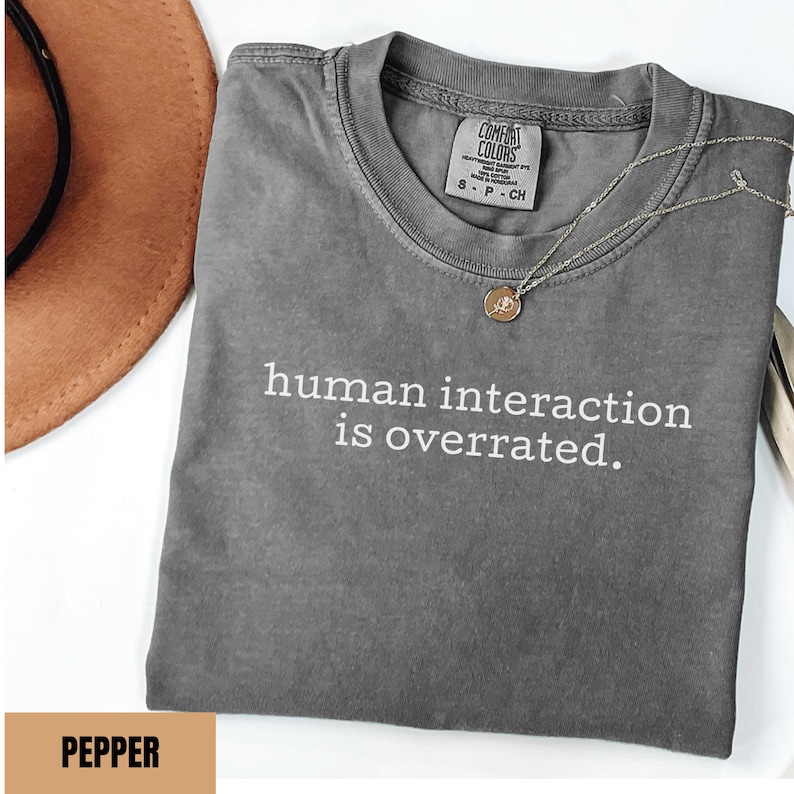 May include: A gray t-shirt with the phrase "human interaction is overrated" in white text. The shirt has a crew neck and short sleeves. A gold necklace with a small pendant is on the shirt. A brown hat is in the background.