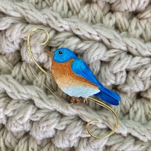 May include: A bluebird brooch with a gold-colored wire perch. The bird has blue, brown, and white painted details. The brooch is set against a textured, off-white knitted background.