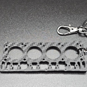 May include: A black and silver keychain featuring a detailed replica of a car engine block. The keychain is attached to a silver split ring and a silver chain.