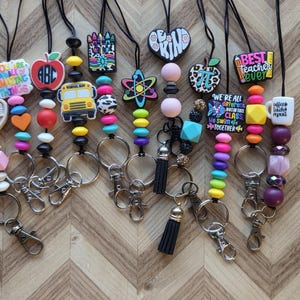 May include: A collection of ten lanyards featuring colorful silicone beads and various school-themed charms including a yellow school bus, an apple, a crayon box, and text-based pendants like Best Teacher Ever and We Are All Different.