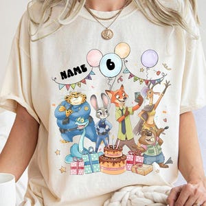 May include: Cream-coloured t-shirt with a vibrant Zootopia-themed birthday graphic. The design includes characters, balloons, a cake, presents, and the number 6. The word "NAME" is also displayed.
