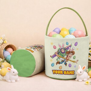 Puede incluir: Una cesta de Pascua verde claro con un asa verde, llena de huevos coloridos. La cesta presenta un gr&aacute;fico de dibujos animados de Buzz Lightyear y Woody. El texto "YOUR NAME" est&aacute; impreso en la parte delantera. Un recipiente cil&iacute;ndrico a juego est&aacute; al lado de la cesta.