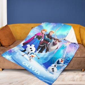 Puede incluir: Una manta azul con personajes de la pel&iacute;cula animada "Frozen", incluyendo a Anna, Elsa, Kristoff, Olaf y Sven. La manta tiene un fondo nevado con el nombre "Olivia" impreso.