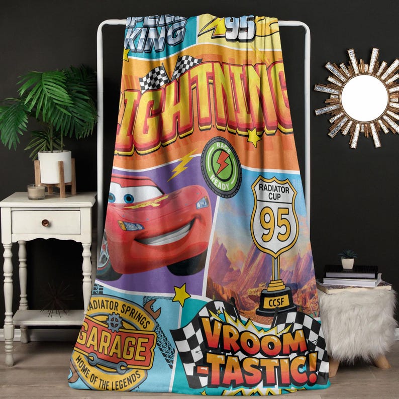 May include: A colorful blanket featuring Lightning McQueen from the movie Cars. The design includes the words "Lightning," "Radiator Cup 95," and "Vroomtastic!" in bold, vibrant colors. The blanket is displayed on a white rack.