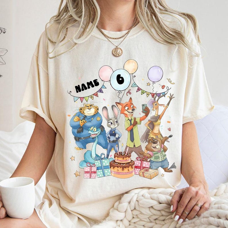 May include: Cream-coloured t-shirt with a vibrant Zootopia-themed birthday graphic. The design includes characters, balloons, a cake, presents, and the number 6. The word "NAME" is also displayed.