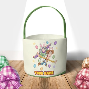 Puede incluir: Cesta de Pascua de color crema con asa y ribete verdes. La cesta presenta un gr&aacute;fico colorido de Buzz Lightyear y Woody, ambos con orejas de conejo, rodeados de huevos de Pascua y estrellas. El texto "YOUR NAME" est&aacute; en la parte inferior.