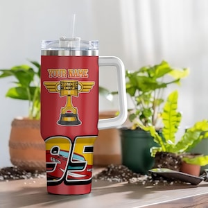 Puede incluir: Un vaso t&eacute;rmico rojo con asa y tapa blancas. El vaso presenta un dise&ntilde;o "YOUR NAME" sobre un trofeo Piston Cup dorado y el n&uacute;mero 95 con un gr&aacute;fico de coche de carreras rojo. Incluye una pajita transparente.