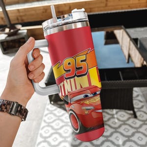 Puede incluir: Vaso t&eacute;rmico rojo con Lightning McQueen de la pel&iacute;cula Cars. El vaso tiene un asa blanca, una tapa transparente y una pajita. El dise&ntilde;o incluye el n&uacute;mero 95 y la palabra "Lightning".