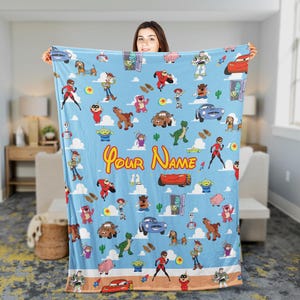 May include: A light blue blanket featuring a repeating pattern of cartoon characters, including a red car, a green dinosaur, and a cowboy. The blanket has the text "Your Name" in yellow script.