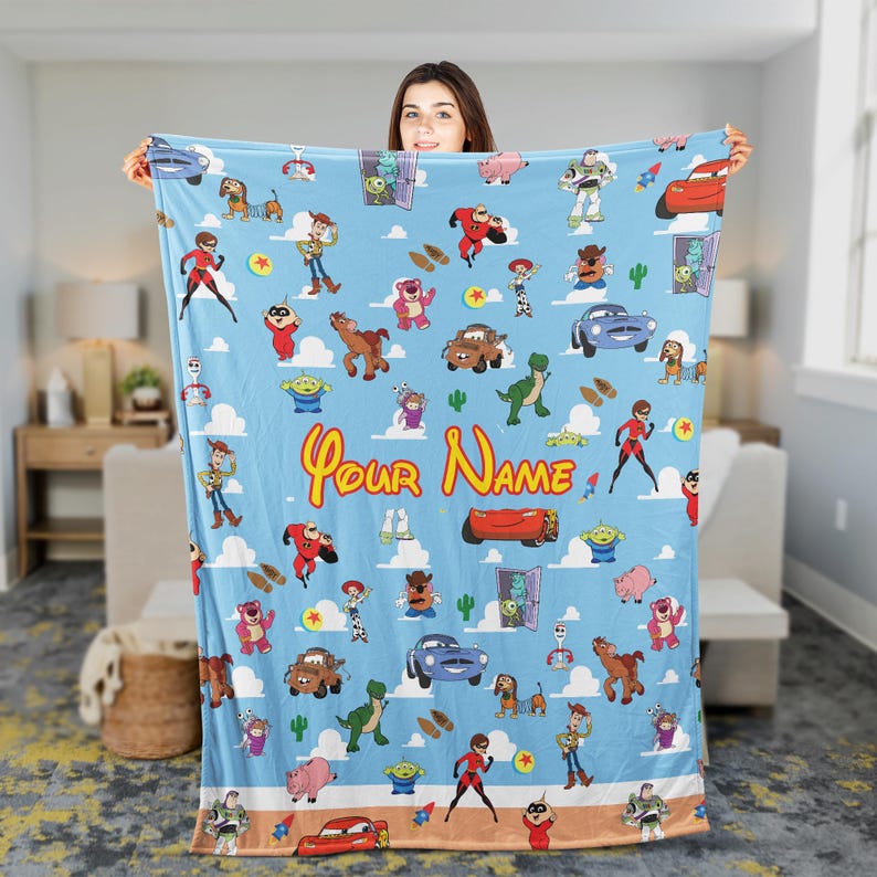 May include: A light blue blanket featuring a repeating pattern of cartoon characters, including a red car, a green dinosaur, and a cowboy. The blanket has the text "Your Name" in yellow script.