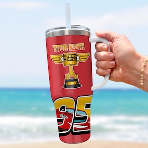Puede incluir: Vaso rojo con asa y pajita blancas. El vaso presenta el texto "YOUR NAME" sobre un gr&aacute;fico de trofeo Piston Cup dorado. Debajo hay un gr&aacute;fico del n&uacute;mero 95 y un coche de carreras rojo.