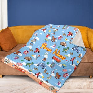 May include: A light blue blanket featuring cartoon characters and the text "Your Name". The blanket has a white sherpa lining and is draped over a brown sofa. The blanket is decorated with various cartoon characters.
