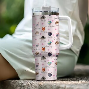 Puede incluir: Un vaso de acero inoxidable con asa y tapa blancas. Presenta un fondo rosa con caras de animales de dibujos animados y texto. El vaso incluye una pajita y est&aacute; dise&ntilde;ado para bebidas fr&iacute;as.