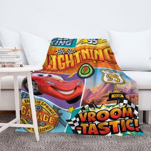 May include: A colorful blanket featuring Lightning McQueen from the Cars movie. The design includes the text "Lightning 95", "Race Ready", and "Vroom Tastic!" in a vibrant, cartoon style. The blanket is draped over a white couch.
