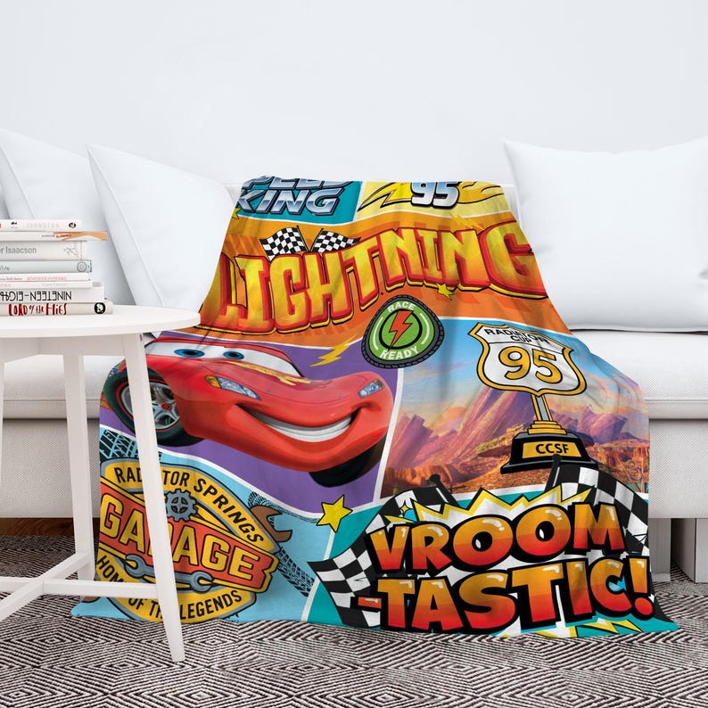 May include: A colorful blanket featuring Lightning McQueen from the Cars movie. The design includes the text "Lightning 95", "Race Ready", and "Vroom Tastic!" in a vibrant, cartoon style. The blanket is draped over a white couch.