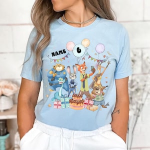May include: Pale blue t-shirt with a vibrant Zootropolis-themed birthday graphic. The design includes cartoon characters, a cake, presents, balloons, and the number 6. The word "NAME" is also displayed.