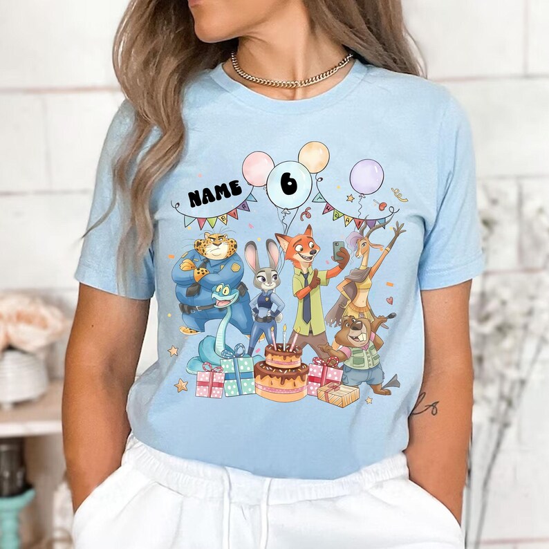 May include: Pale blue t-shirt with a vibrant Zootropolis-themed birthday graphic. The design includes cartoon characters, a cake, presents, balloons, and the number 6. The word "NAME" is also displayed.